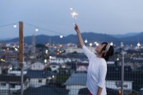 Smiling young Japanese woman holding sparkler on rooftop in urban setting. — Stock Photo
