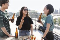 Jóvenes hombres y mujeres japoneses de pie en la azotea en un entorno urbano, bebiendo cerveza con aperitivos . - foto de stock