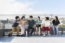 Улыбающаяся группа молодых японских мужчин и женщин, сидящих с бутылками пива на крыше в городской обстановке . — стоковое фото