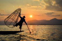Pescador balanceando em uma perna em um barco, segurando uma cesta de pesca grande, pesca da maneira tradicional no Lago Inle ao pôr do sol, Mianmar. — Fotografia de Stock