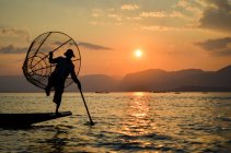 Традиционный рыбак балансирует на одной ноге на лодке, держа в руках рыболовную корзину, ловит рыбу на озере Инле на закате, Мьянма. — стоковое фото