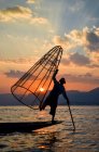Tradicional pescador balanceamento em uma perna em um barco, segurando cesta de pesca, pesca no Lago Inle ao pôr do sol, Mianmar. — Fotografia de Stock