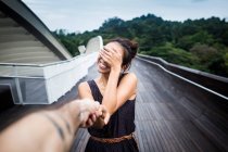 Smiling young woman standing on a bridge, covering her face, holding male hand. — Stock Photo
