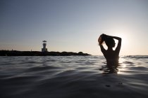 Visão traseira da mulher tomando banho no oceano ao pôr do sol, farol à distância. — Fotografia de Stock