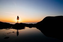 Silhueta de mulher em pé sobre uma rocha junto ao oceano ao pôr do sol. — Fotografia de Stock