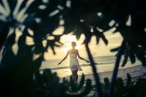 Вид спереду молода жінка, що йде вздовж пляжу на заході сонця, пальми на передньому плані . — стокове фото