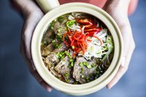 Alto ângulo perto de mãos segurando tigela com sopa asiática contendo arroz vermicelli, carne bovina e chili guarnição . — Fotografia de Stock