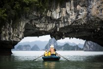 Visão traseira do homem usando chapéu de palha transportando pequeno grupo de pessoas em barco, remo sob arco de rocha natural, Bai Tu Long, Vietnã . — Fotografia de Stock
