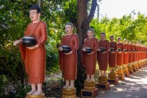 Ряд статуй буддийских монахов с красными одеждами и чашами с подаянием в саду буддийского храма в Сиемреапе, Камбоджа — стоковое фото