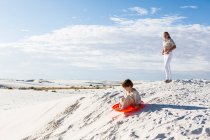 Дети играют в пейзаже песчаных дюн, один на оранжевых санках. — стоковое фото