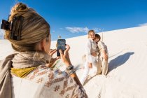 A woman taking picture of her children with a smart phone in white sand dunes landscape under blue sky. — Stock Photo