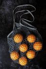 High angle close up of oranges in grey net bag on black background. — Stock Photo