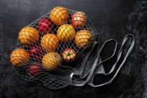 Hohe Nahaufnahme von roten Äpfeln und Orangen in grauer Netztasche auf schwarzem Hintergrund. — Stockfoto