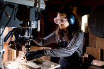 Frau mit langen braunen Haaren mit Latzhose, Schutzbrille und Ohrenschutz steht in Holzwerkstatt mit Bohrmaschine. — Stockfoto