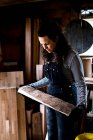Woman with long brown hair wearing dungarees standing in wood workshop, holding piece of wood. — Stock Photo
