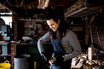 Woman with long brown hair wearing dungarees, working in wood workshop. — Stock Photo