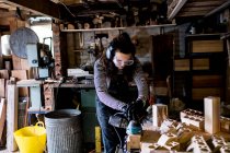 Женщина с длинными каштановыми волосами в балахонах, защитных очках и ушных протекторах, стоящая в деревообрабатывающей мастерской, с использованием сандера. — стоковое фото