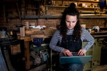 Woman with long brown hair wearing dungarees standing in wood workshop, typing on laptop computer. — Stock Photo