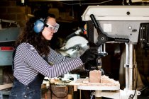 Frau mit langen braunen Haaren mit Latzhose, Schutzbrille und Ohrenschutz steht in Holzwerkstatt mit Bohrmaschine. — Stockfoto