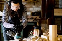 Женщина с длинными каштановыми волосами в балахонах, защитных очках и ушных протекторах, стоящая в деревообрабатывающей мастерской, с использованием сандера. — стоковое фото
