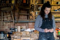 Женщина с длинными каштановыми волосами в балаклавах стоит в мастерской по дереву, пользуясь мобильным телефоном. — стоковое фото