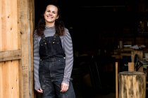 Woman with long brown hair wearing dungarees standing at entrance to wood workshop, smiling at camera. — Stock Photo