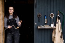Женщина с длинными каштановыми волосами в балаклавах стоит у входа в мастерскую деревообработки, глядя в камеру. — стоковое фото