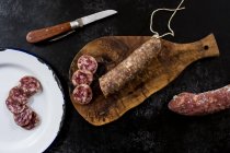 High angle close up of knife, sliced salami on wooden cutting board and white enamel plate on black background. — Stock Photo