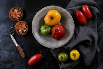 High angle close up of knife, grey plate and cloth and a selection of fresh tomatoes on black background. — Stock Photo