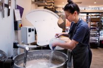 Woman wearing apron standing in an artisan bakery, pouring water into industrial mixer. — Stock Photo