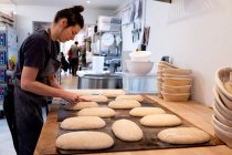 Woman wearing apron standing in an artisan bakery, shaping sourdough loaves for baking. — Stock Photo