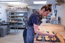 Femme portant un tablier debout dans une boulangerie artisanale, brossant des petits pains à la cannelle sur une plaque à pâtisserie . — Photo de stock