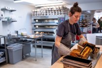 Femme portant un tablier et des gants de four debout dans une boulangerie artisanale, se révélant des pains fraîchement cuits de pain semé . — Photo de stock
