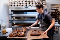 Frau mit Schürze steht in einer handwerklichen Bäckerei und legt frisch gebackene Brote auf Holzbretter. — Stockfoto