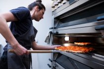 Donna che indossa grembiule in piedi in una panetteria artigianale, mettendo la pizza in forno . — Foto stock