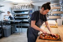 Woman wearing apron standing in an artisan bakery, slicing freshly baked pizza. — Stock Photo