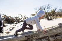 Garçon de six ans sur une plage grimpant dans un tronc d'arbre de bois flotté — Photo de stock