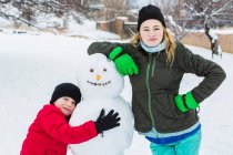 Brother and sister, a young boy and teenage girl leaning on snowman in winter — Stock Photo