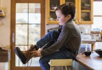 Six year old boy putting his boots on, sitting on a high stool. — Stock Photo