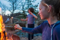 Teenage girl making smores with her brother over a fire in a garden at dusk. — Stock Photo