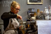 Blond woman wearing glasses and apron standing at espresso machine in a cafe, pouring milk into metal jug. — Stock Photo