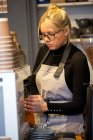 Блондинка в очках и фартуке, стоящая у кофеварки в кафе, пенящая молоко. — стоковое фото
