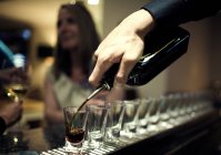 Крупним планом бармен наливає напої з пляшки в ряд дробових окулярів, що стоять на барній стійці, жінка сидить на фоні . — стокове фото