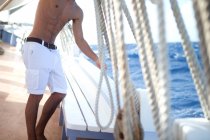 Uomo senza maglietta che indossa pantaloncini bianchi in piedi sul ponte di una barca a vela, sartiame. — Foto stock