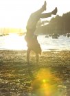 Homme faisant un stand de main sur le rivage, la mer et amarrés voiliers en arrière-plan, la lumière du soleil. — Photo de stock