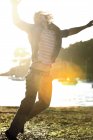 Junger Mann springt auf einen Kieselstrand, Arme erhoben, im Hintergrund festgemachte Segelboote, Sonnenlicht. — Stockfoto