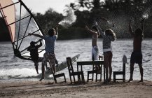 Small group of men and women standing on a sandy beach around table and chairs, arms raised, holding bottles, watching a windsurfer. — Stock Photo