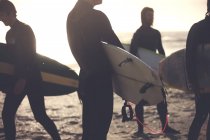 Quatre hommes portant des combinaisons debout sur une plage de sable, portant des planches de surf. — Photo de stock