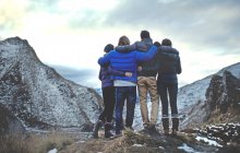 Rückansicht von vier Menschen, die Arm in Arm auf einem Berg stehen, in der Ferne schneebedeckte Gipfel. — Stockfoto