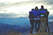 Вид сзади на четыре человека, стоящих рука об руку на горе, снежные вершины вдалеке. — стоковое фото